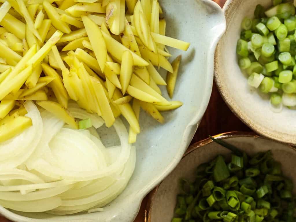 Close-up of a bowl containing julienned potato, another with sliced onions, and a third with chopped green onions. The ingredients are fresh and vibrant, displayed in ceramic bowls on a wooden surface.